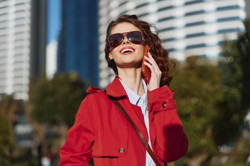 Young woman with curly hair wearing a red coat and sunglasses smiles while talking on a smartphone outdoors in an urban environment on a sunny day.