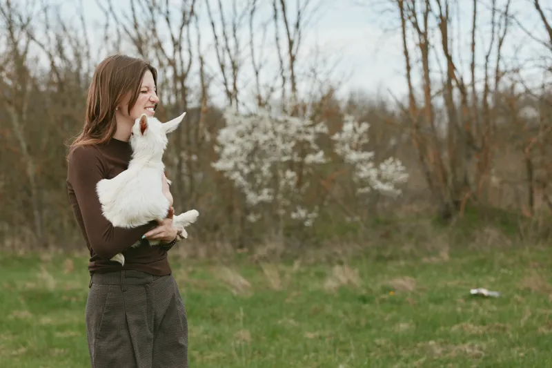 Woman outdoors in a park holds a playful goat kid, sharing a kiss as the affectionate animal nuzzles her, conveying joy and trust. Bright natural light, casual attire, rustic scenery, and a warm