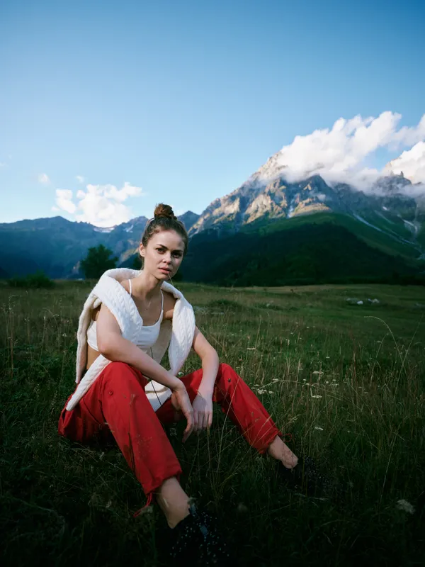Outdoor scene with a woman seated in a grassy meadow and mountains in the background, wearing a white top red pants and a light knit shawl, capturing a peaceful nature moment