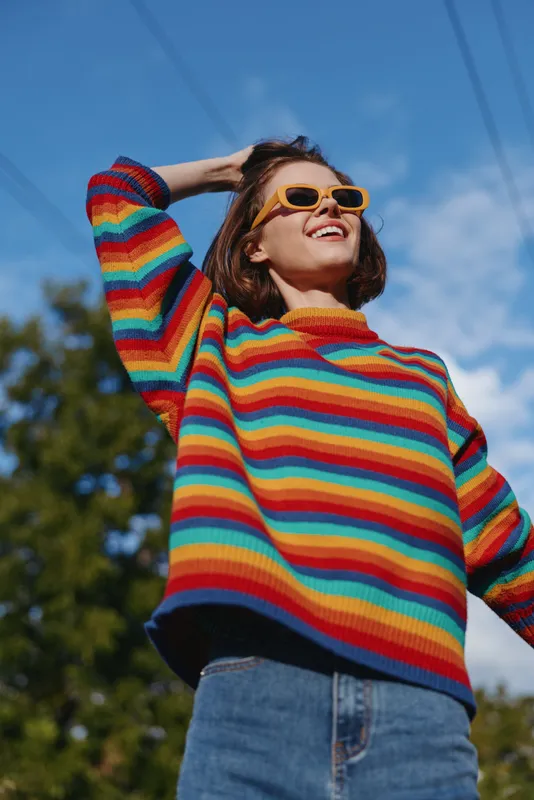 woman in a rainbow sweater wearing yellow sunglasses smiling outdoors, casual outfit with jeans, brunette in her 20s leaning back and enjoying sunlit blue sky and leafy park atmosphere