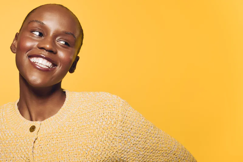Smiling young black woman with dark skin wearing a yellow knitted sweater looks away happily against a bright yellow background. Concept of joy, fashion, and positive mood.