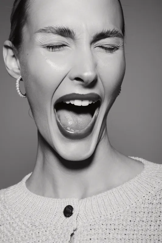 Young Caucasian woman with fair skin in knitted sweater screams with closed eyes in black and white portrait.