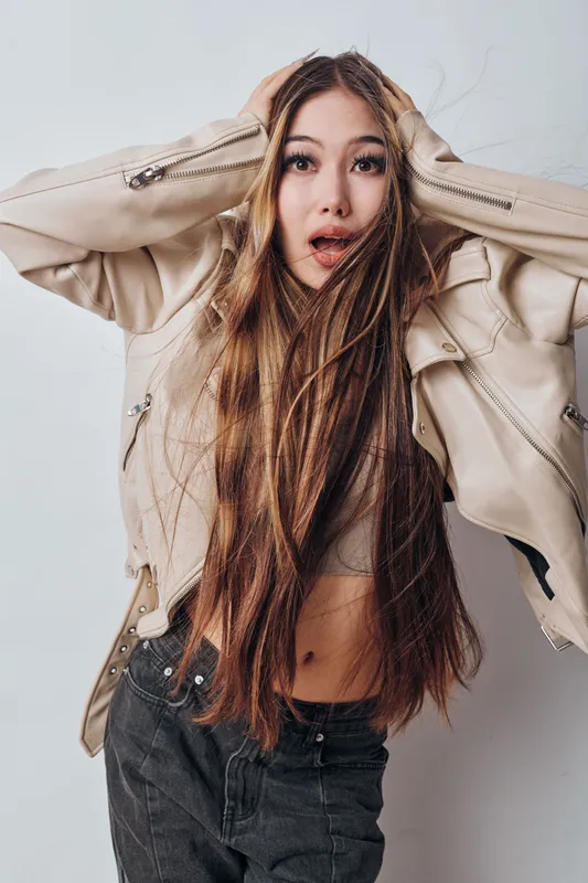 Young woman with long hair wearing light jacket and dark pants looks surprised with hands on head against light background. Expressive emotion, casual style, modern fashion portrait.