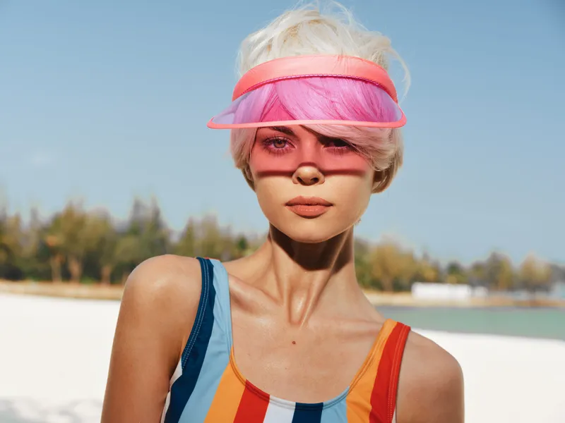Close up portrait of a young woman at a sunny beach wearing a vibrant striped swimsuit and a translucent pink visor, with luminous skin and relaxed expression.