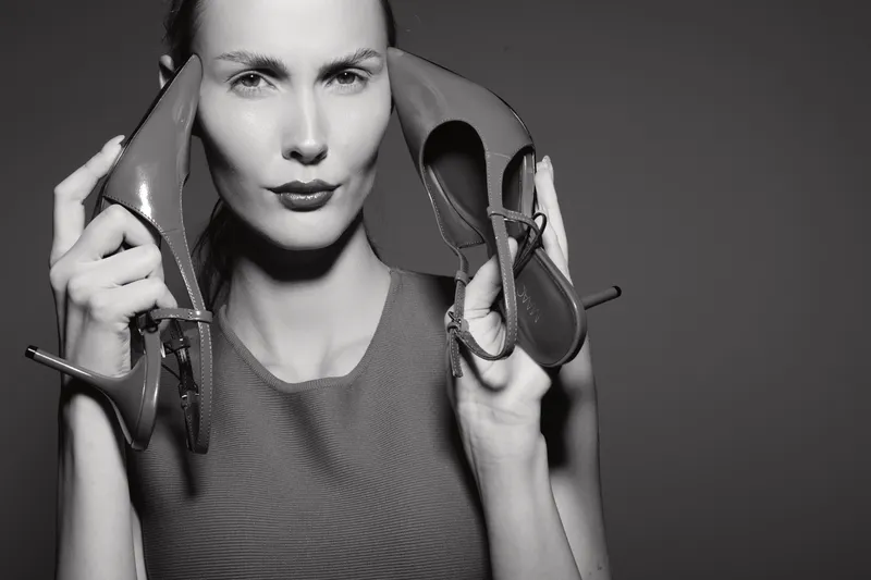 White woman with light skin holds high heel shoes near face in studio. Portrait with confident expression on grey background.