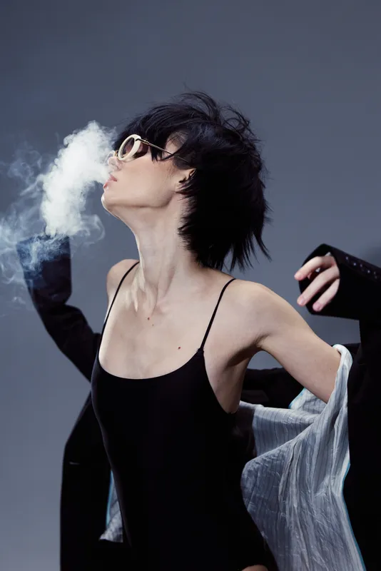 Stylish woman exhaling smoke wearing sunglasses and black outfit in moody lighting