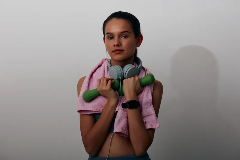 Woman in athletic wear with pink towel, headphones and green dumbbells, posing in a studio for fitness and sport training with a focused expression.
