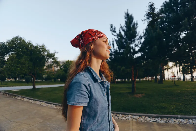 Lifestyle woman in modern boho-western denim street style wearing a red bandana outdoors, natural light, film color, casual confident look in urban park setting.