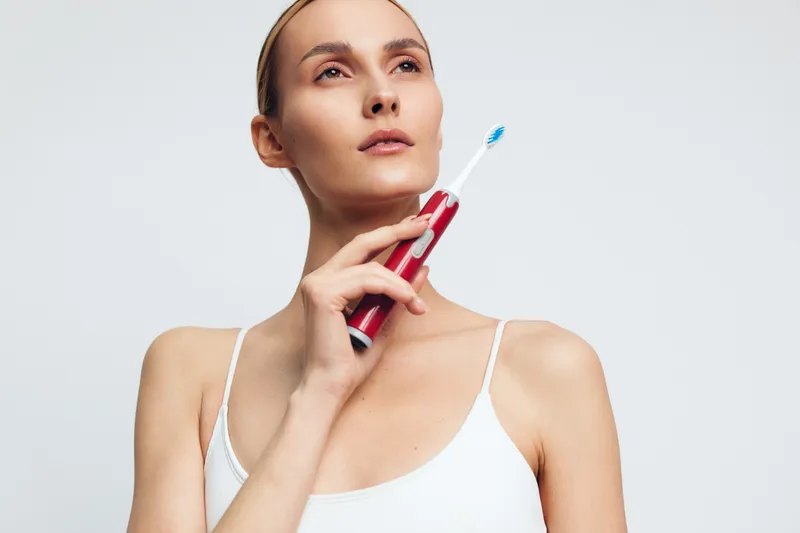 Confident woman holds a red toothbrush under bright light, looking upward with a calm smile, wearing a white sleeveless top, isolated on a pale background, promoting personal dental care.
