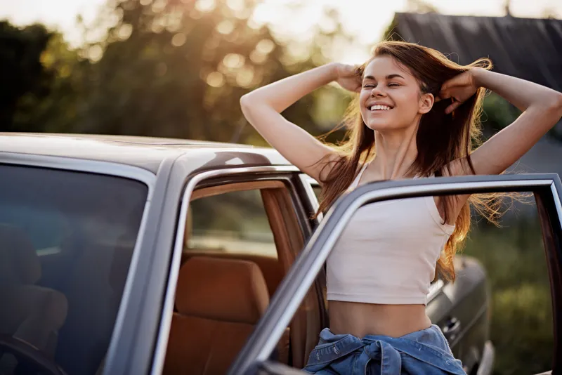 Young woman enjoying summer sunlight outside a vintage car, embodying freedom and joy with wind in her hair, perfect for lifestyle and travel themes.