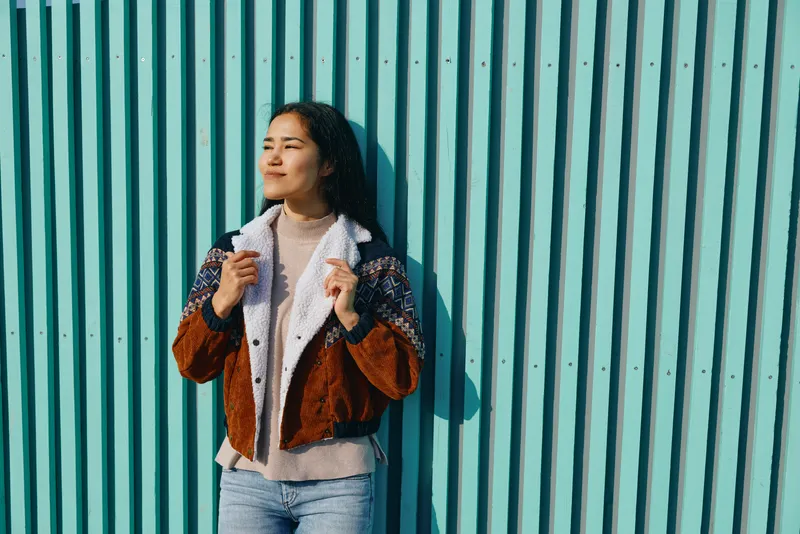 Woman wearing a brown shearling jacket and denim jeans stands against a teal corrugated wall, adjusting her collar with a confident gaze, outdoor fashion, urban style, casual chic and sunny mood