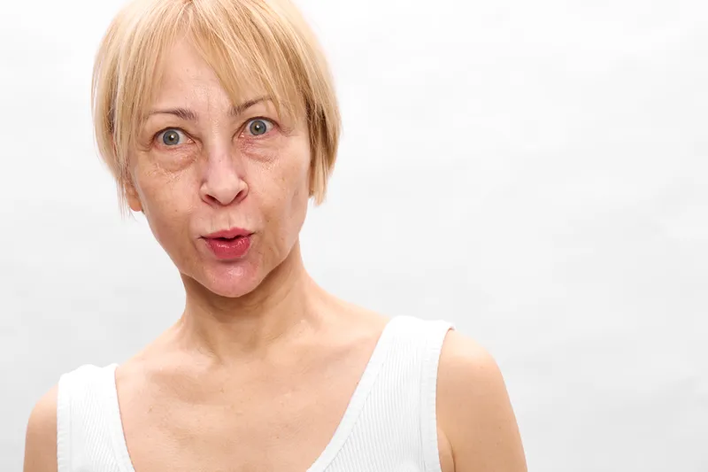 Portrait of a worried woman with short blond hair, wide eyes and open mouth, wearing a light white sleeveless top against a clean white background, emotional stress concept