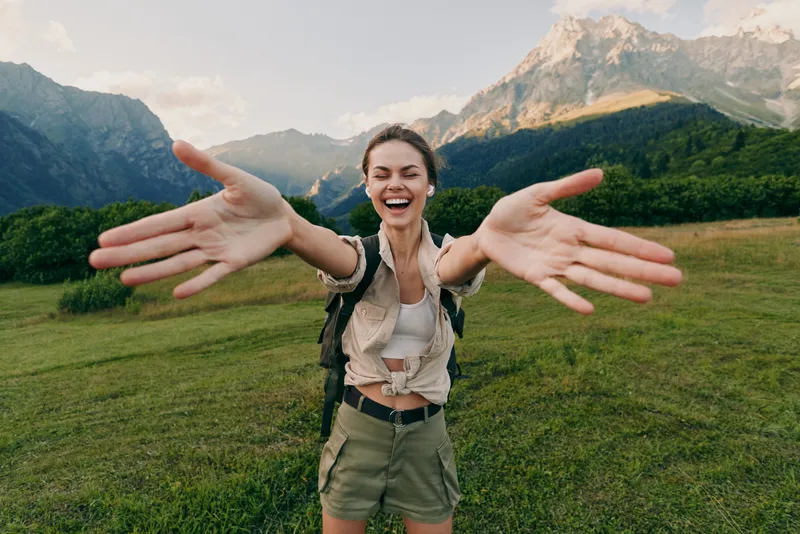 Young woman with a backpack joyfully reaches out with open arms in a green meadow with mountains in the background during a sunny day.