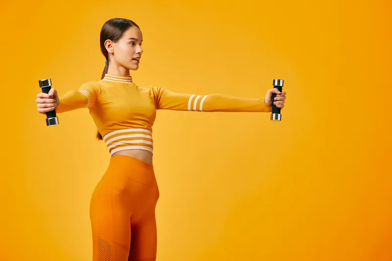 Fitness woman in bright orange activewear holds dumbbells in a lateral raise against a vivid orange backdrop, showcasing strength, balance and athletic training.