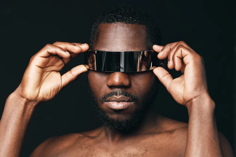African man with dark skin wearing futuristic visor glasses in creative conceptual visual style. Portrait of confident male model with bare chest and modern accessory on black background.