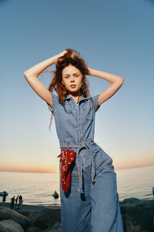 lifestyle woman in modern boho-western denim jumpsuit with red bandana posing at sunset beach, film color, casual street style, relaxed mood, scenic ocean background