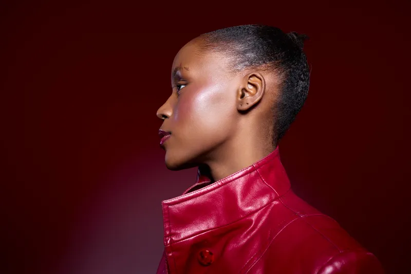 Profile of a young African woman with dark skin wearing a red leather jacket. The studio lighting highlights her smooth skin and elegant features against a dark red background.
