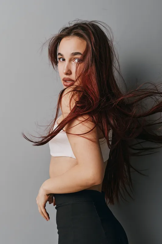 Young woman with long flowing hair posing in stylish outfit against gray background. Fashion model with natural makeup looking at camera with confident expression and casual posture.