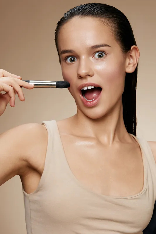 Young woman with wet hair applying makeup, displaying excitement and confidence in a neutral toned background, perfect for beauty and cosmetic themes