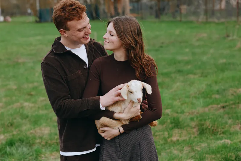 Couple, woman, man, goat, kid, outdoors, lifestyle, affection, animals, romantic. A smiling pair in a grassy park cradle a gentle goat kid, capturing a warm candid moment of tenderness and connection