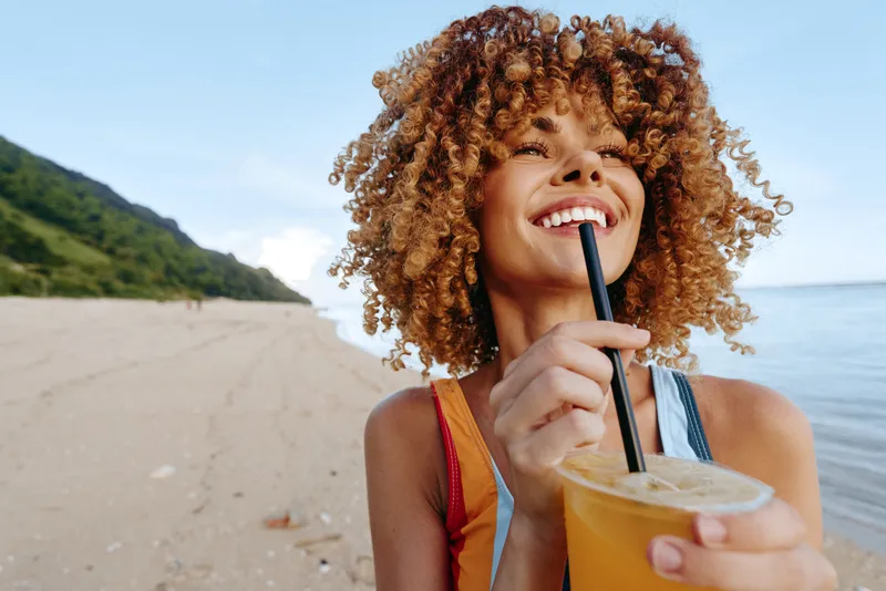 Woman with curly hair laughing outdoors, holding a straw drink in a clear cup near the sea, sunny beach background and green hills, soft natural light, summer lifestyle, refreshment, joy, casual