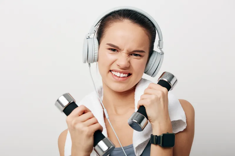 Woman fitness sport enthusiast lifts dumbbells with headphones and towel, showing determination during a dynamic home workout for strength and endurance.