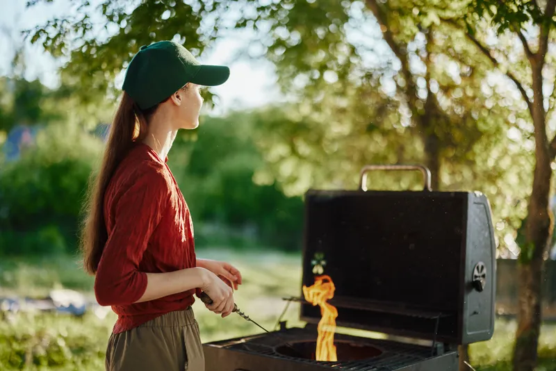 Young woman in a green cap grilling outdoors in a backyard during summer, enjoying the vibrant atmosphere with flames and greenery
