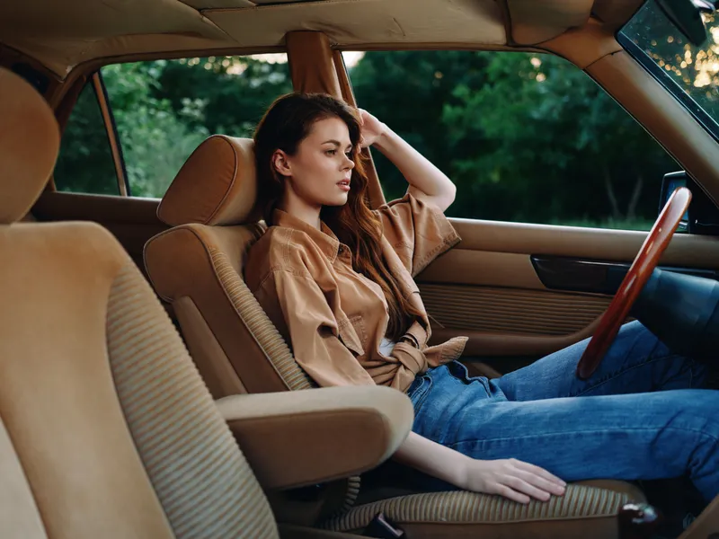 Thoughtful young woman sitting in a vintage car, dressed in casual brown shirt and jeans, reflecting in a serene environment, perfect for seasonal lifestyle imagery.