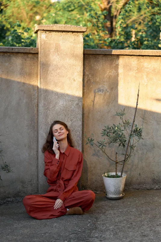 Relaxed woman in a red jumpsuit sits against a sunlit outdoor wall, conveying lifestyle and wellbeing, calm reflection, and quiet moments of contemplation in warm light, natural textures, simple