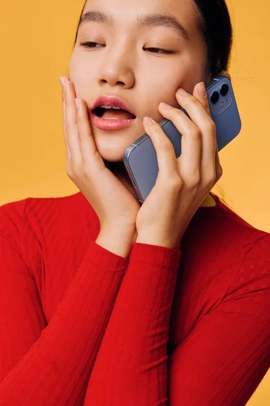 Young woman with long dark hair in red sweater talking on smartphone against yellow background, expressing surprise or shock with open mouth and hand on cheek closeup portrait