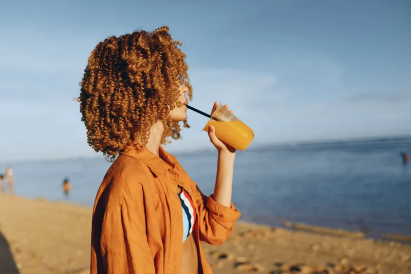 Woman with curly hair at the beach, ocean summer sunlight, drinking from a bottle, relaxed posture, calm travel mood, softly blurred waves behind, natural colors