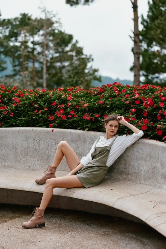 Young woman in green overalls poses confidently among vibrant red flowers, showcasing a relaxed, natural style in an outdoor setting