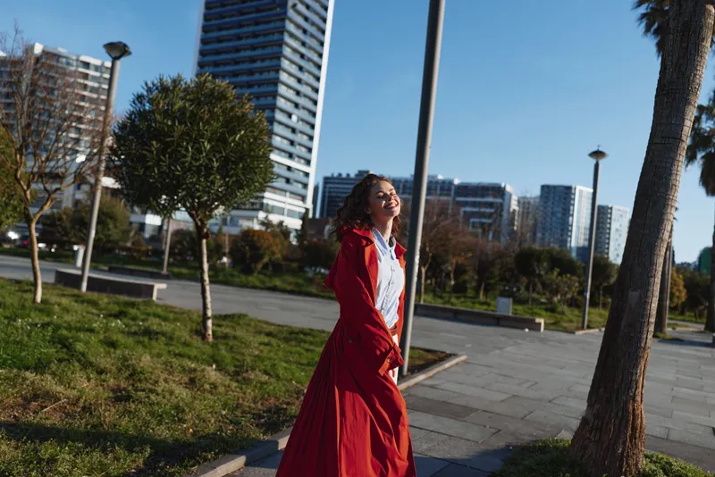 Cheerful woman outdoors, smiling in sunlight, red coat flowing, relaxed walk in a park near modern buildings, clear sky and vivid natural colors