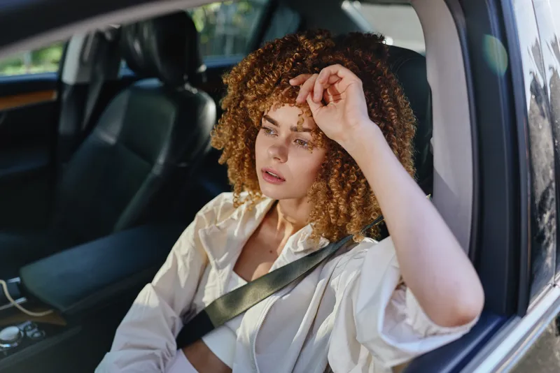 Young woman with curly hair sitting in car wearing seatbelt, thoughtful expression, casual white shirt, natural daylight interior, safety concept.