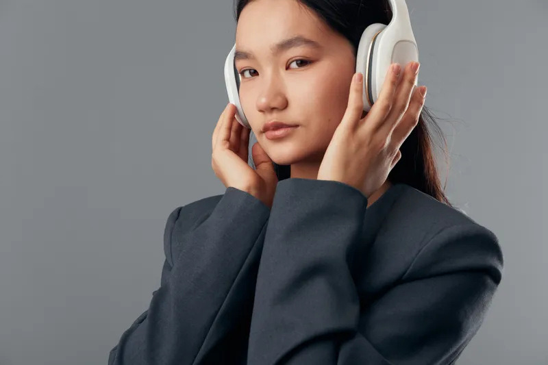 Young woman listens to music with wireless headphones, wearing dark jacket and staring at camera on grey background for portrait and audio technology concept.
