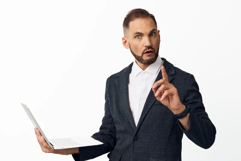 A business professional man stands against an isolated colored background, exuding confidence and surprise while holding a laptop and making a decisive gesture.