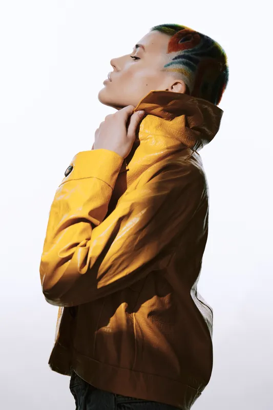 Profile of a stylish woman with colorful shaved hair wearing a mustard yellow jacket, showcasing modern fashion and bold hairstyle in bright studio lighting.
