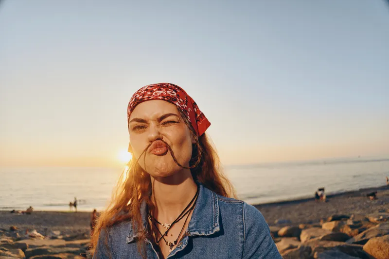 Lifestyle woman in modern boho-western denim street style makes funny face with film color toning at sunset beach background