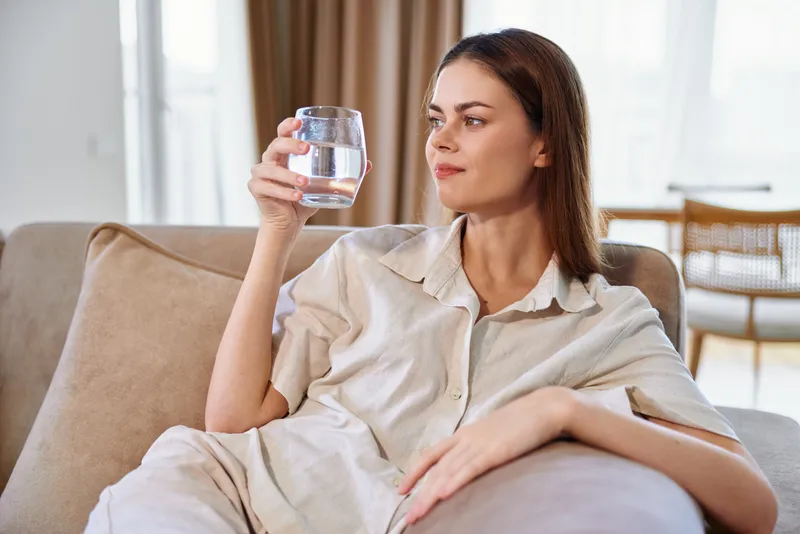 Woman holds a glass of water on a sofa at home, relaxed and thoughtful in casual loungewear, natural sunlight highlighting a hydration moment that evokes wellness and calm domestic lifestyle.