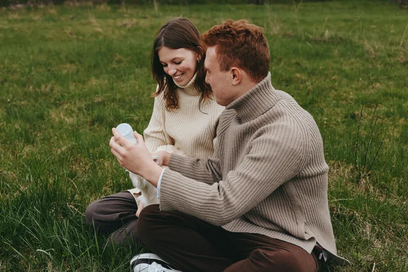 Smiling couple in cozy sweaters sharing a moment with a baby bottle in a grassy field. Natural daylight, warm tones, calm lifestyle scene, affectionate parenting, candid outdoor moment.