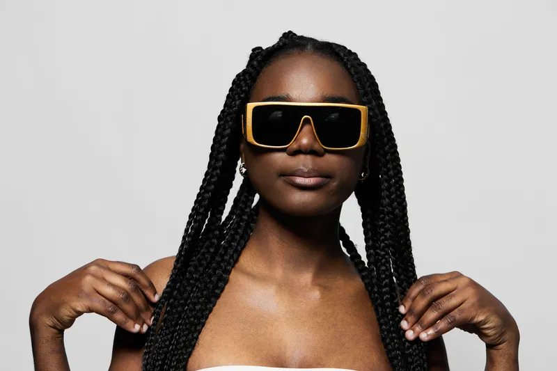 Confident young Black woman with stylish braids wearing oversized sunglasses, exuding summer vibes against a light grey background