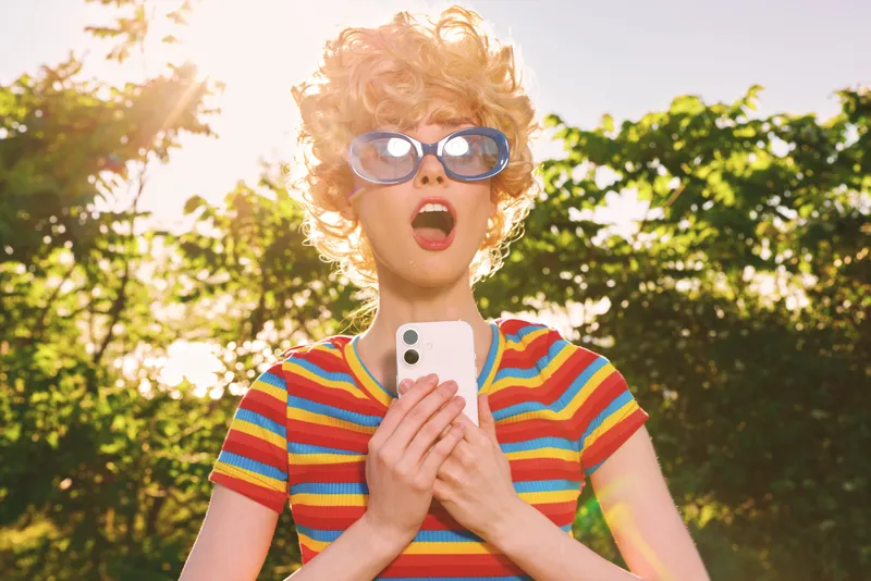 Vibrant outdoor scene shows a surprised woman in a striped shirt and retro sunglasses, clutching a smartphone. A lively moment of spontaneous joy, summer energy, and carefree style.