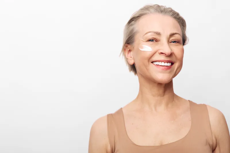 Smiling mature woman with short hair applies white face cream on cheek. Close-up on white background. Concept of skincare, aging beauty, self-care, wellness, and natural glow.