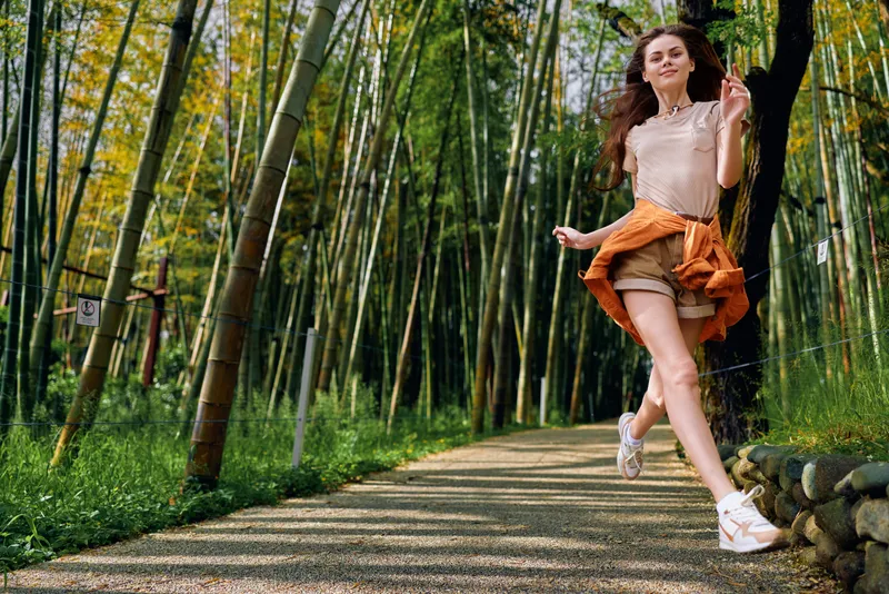 Woman jumping on a forest path through bamboo grove, joyful casual summer outfit with sneakers and light dress, motion and carefree expression during outdoor walk.