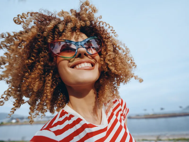 Joyful portrait of a smiling person with curly hair wearing oversized sunglasses and a red striped shirt, captured outdoors on a sunny day with a bright blue sky.