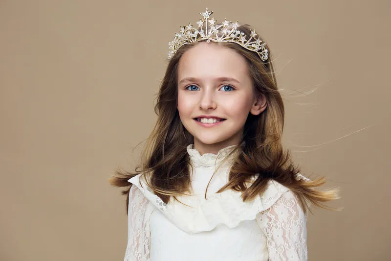 Cute little girl with a crown and a happy smile, wearing an elegant white dress, set against a soft brown background, perfect for seasonal celebrations or childhood themes