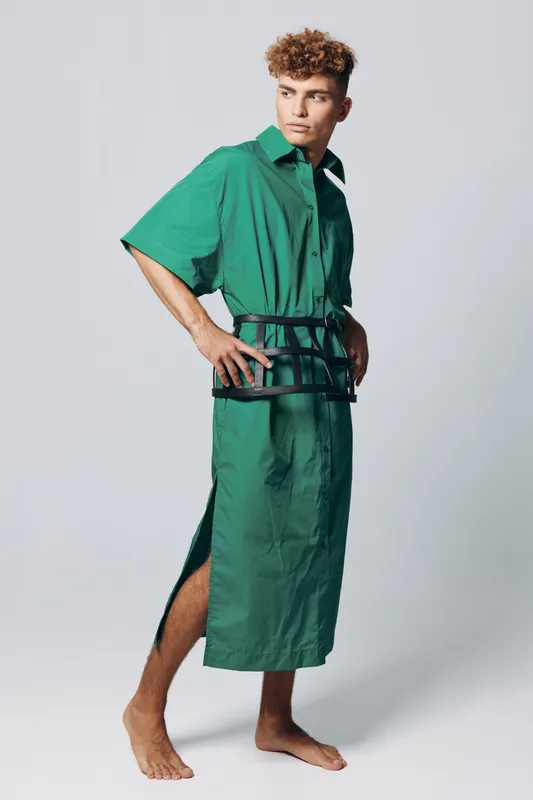 Man in green oversized shirt dress with black belt posing barefoot on white background. Fashion model with curly hair and side slit standing in studio with confident posture.