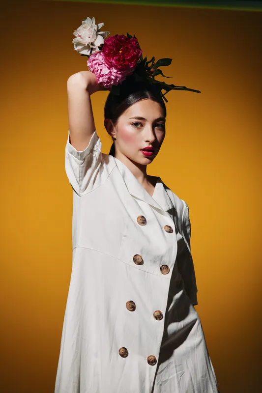 Fashion model with floral hairstyle poses against yellow background. She wears white dress with buttons and bold red lipstick. Creative beauty concept focused on style and elegance.