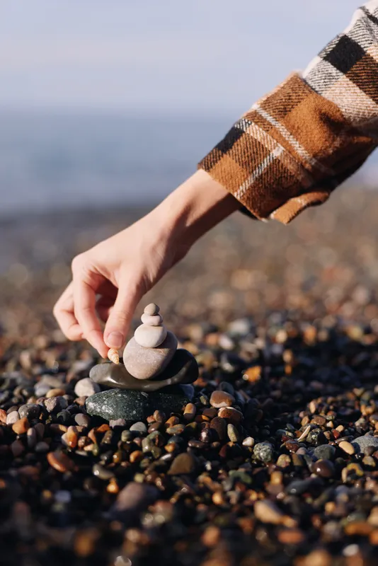 Balance stones on a pebble beach as a hand stacks rocks into a small cairn, a calm seaside moment showcasing texture, focus, and gentle natural light.