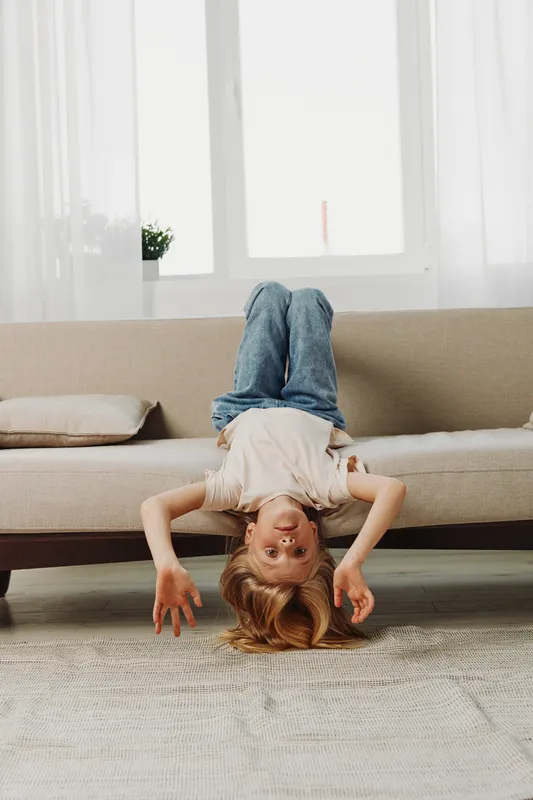 Playful child in casual clothes hangs upside down on beige sofa in bright living room, enjoying carefree moment. Concept of childhood fun, home, and relaxation.